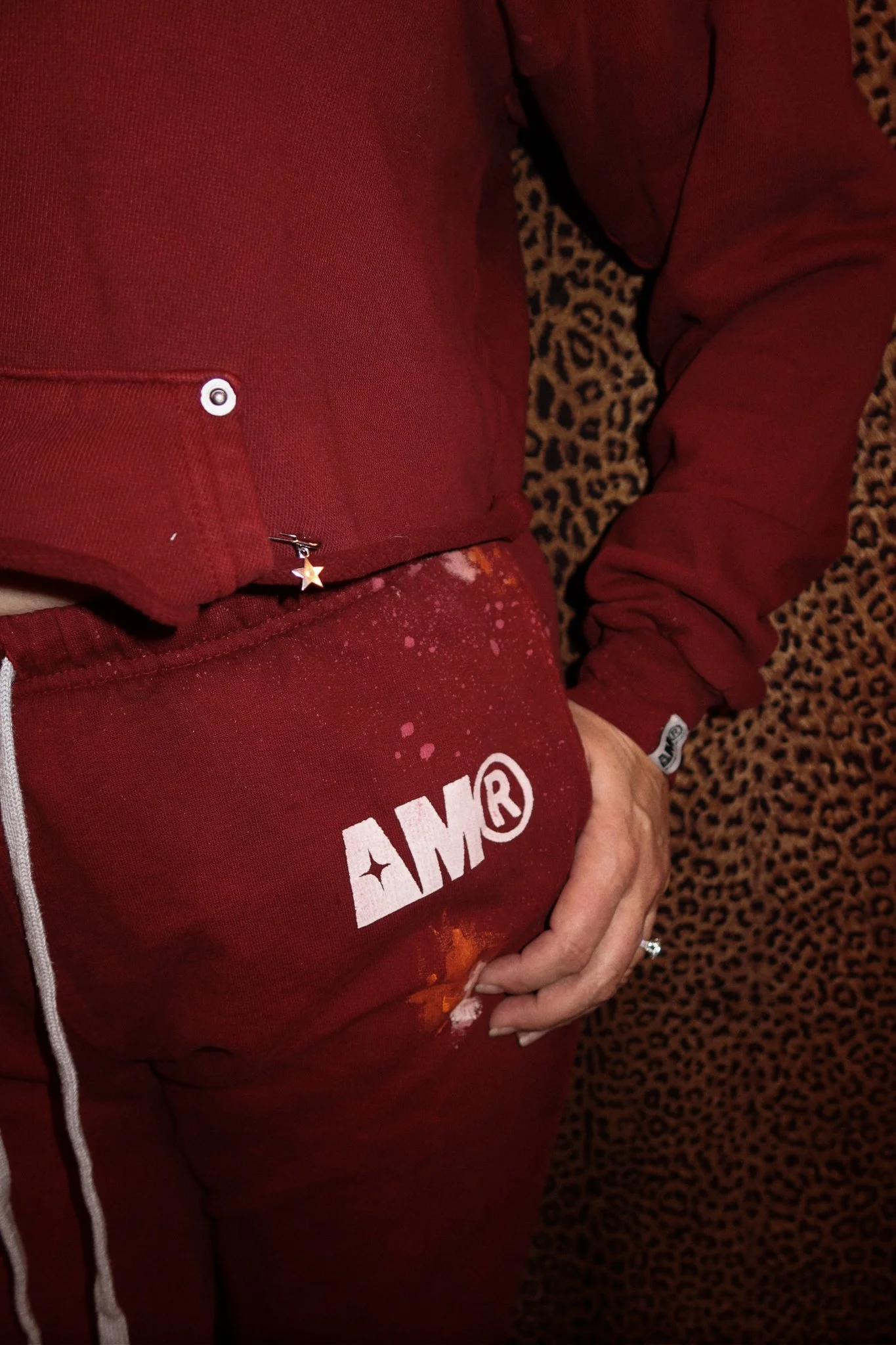 Person wearing a red AMOR sweatshirt and red pants with white paint splatters, standing on a leopard print carpet.