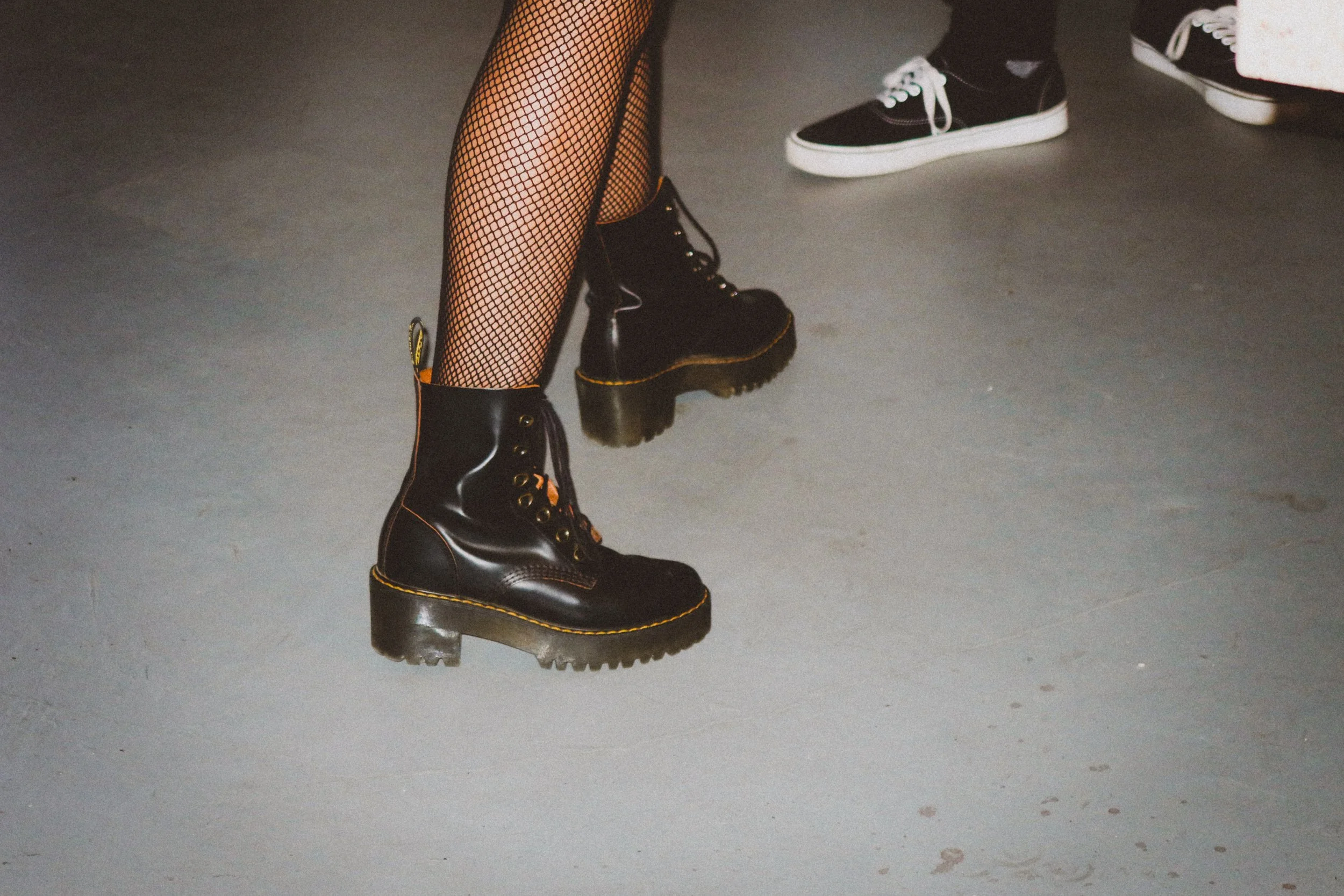 Close-up of a person wearing black combat boots, fishnet stockings, and black pants in a casual setting, with another person wearing black sneakers in the background.