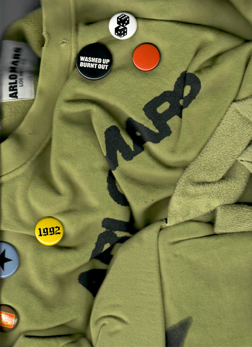 Close-up of a green fabric with various buttons and patches, including a black 'WASHED UP BURNT OUT,' a yellow '1992,' and a few patterned buttons, on a clothing item.
