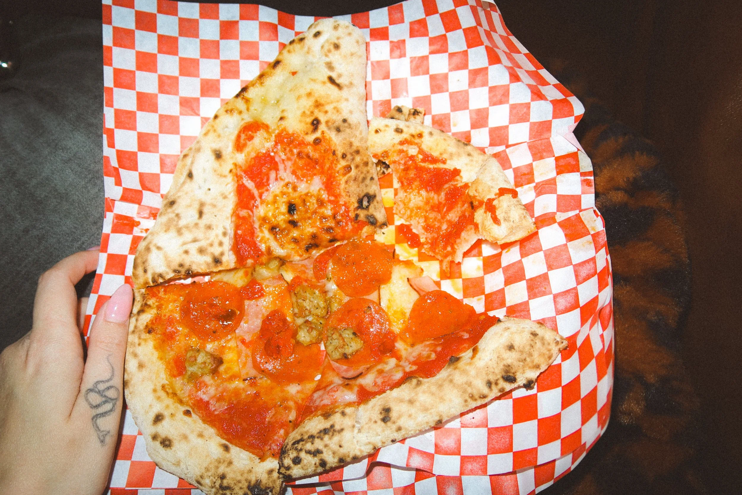 A hand holding a slice of pepperoni pizza on checkered red and white paper with other pizza slices on it.