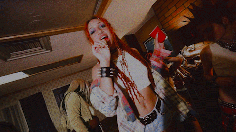 A young woman with red braided hair wearing a plaid shirt and black wristbands, smiling and holding her hand near her face at a social gathering or party.