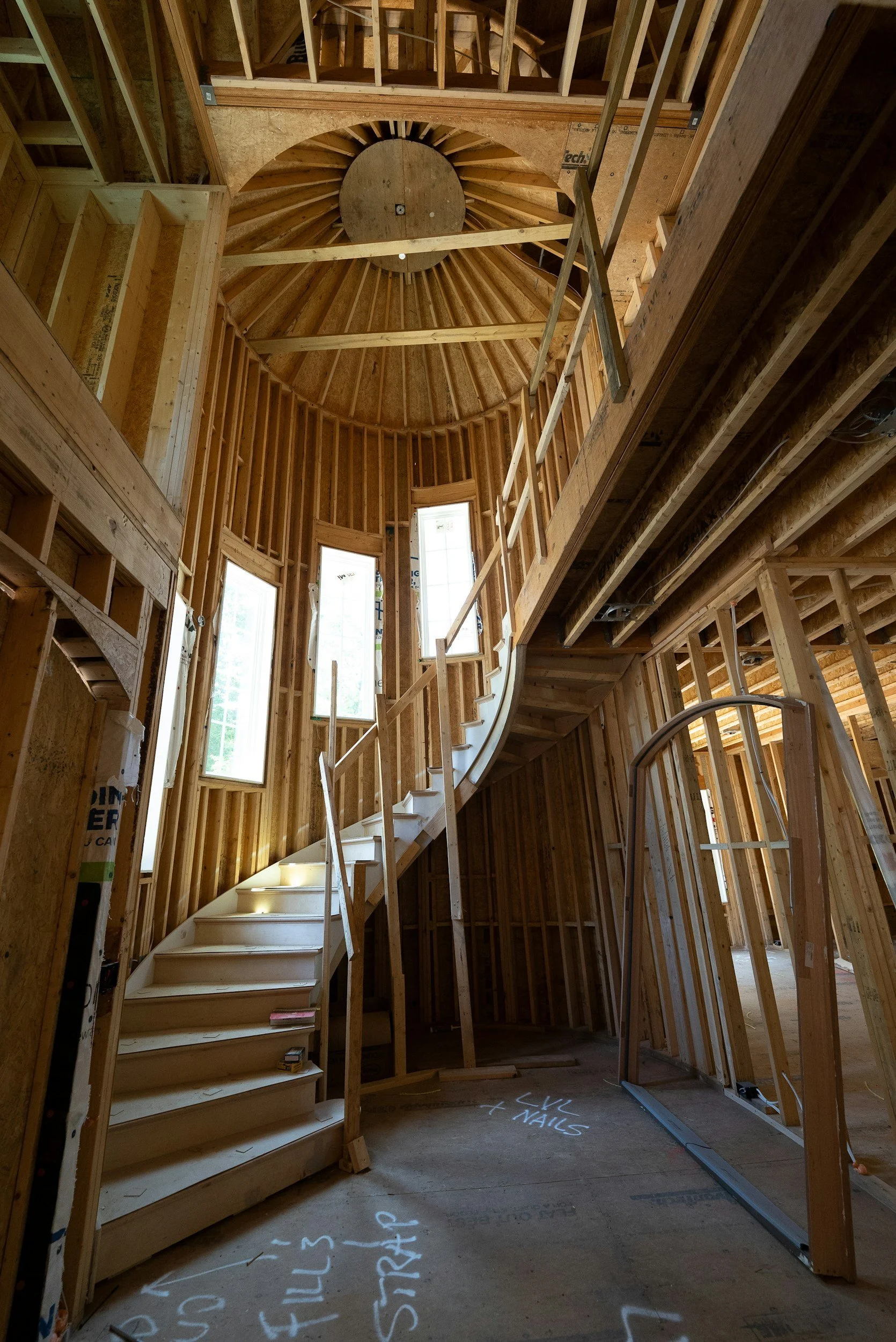Interior view of a house under construction, showcasing a spiral staircase made of wood, surrounded by exposed wooden framing and multiple windows.