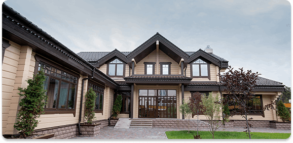 Large modern house with multiple gabled roofs, surrounded by landscaped yard with small trees and bushes.