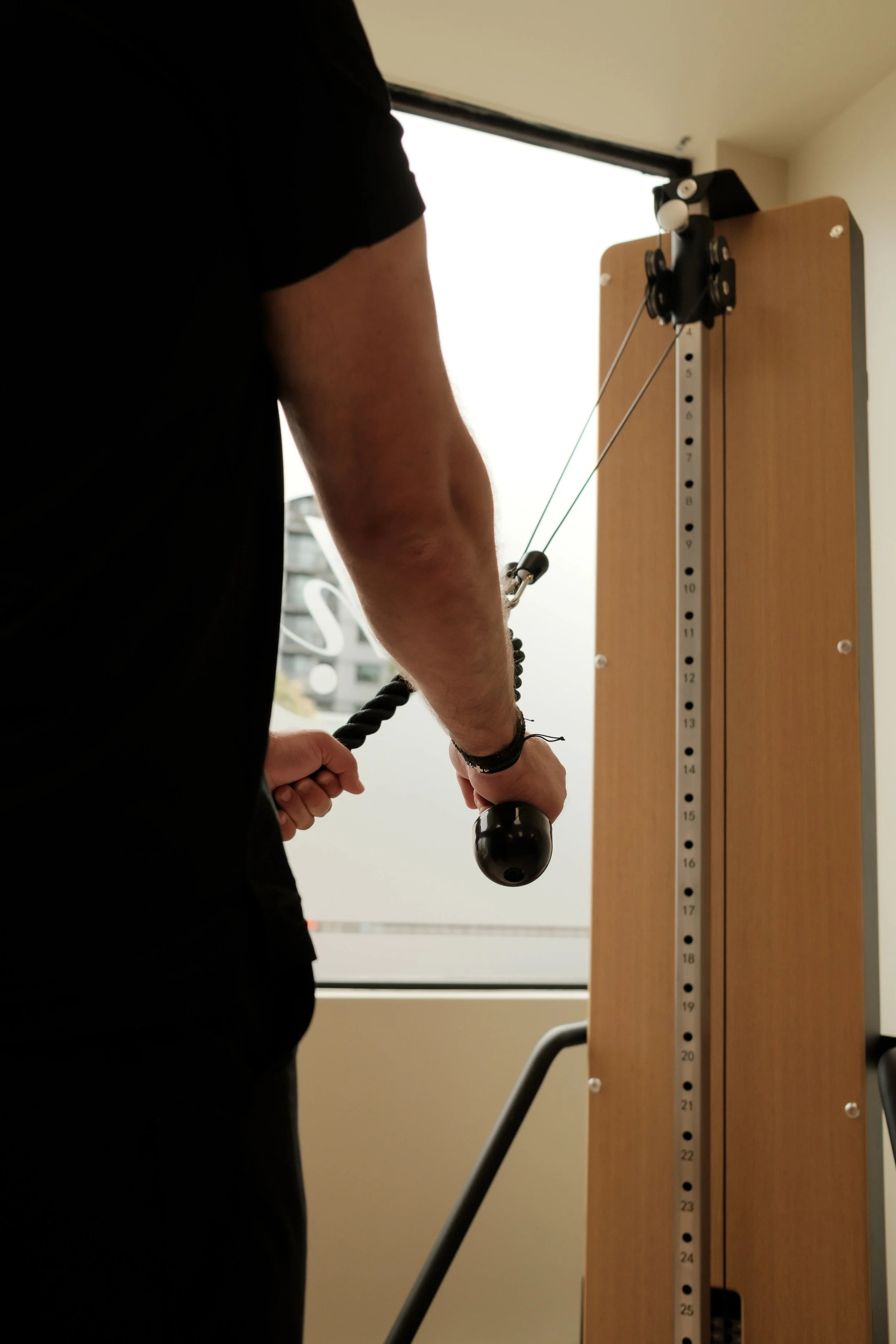 Person using a pulley system for exercise, standing next to a vertically numbered wooden board with cables.