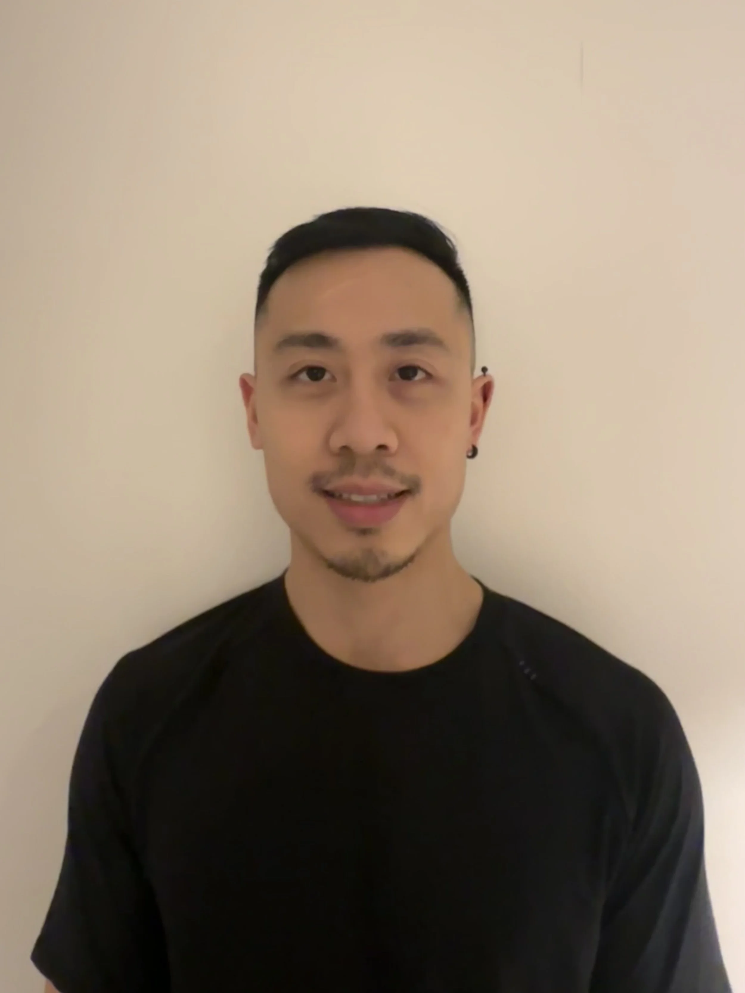 A young man with short black hair, sporting earrings, wearing a black shirt, standing against a plain off-white wall.