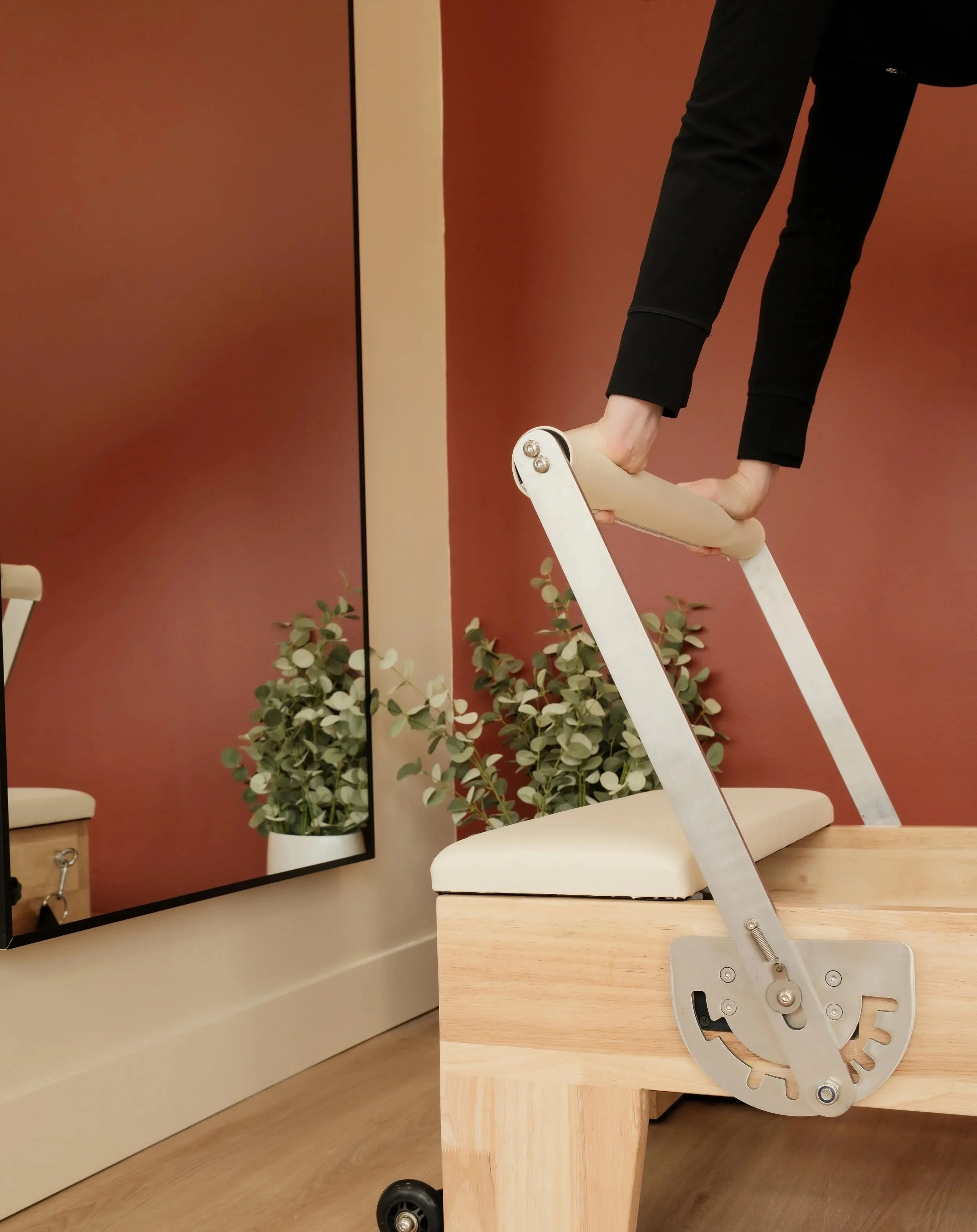 A person holding a folding spatula with a robotic mechanism, standing behind a wooden table in a room with a large mirror, a potted plant, and a white chair.