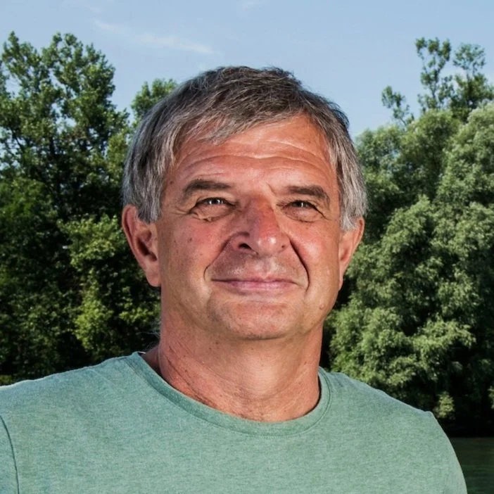 A middle-aged man with gray hair outdoors, smiling slightly, with trees and a river in the background during daytime.