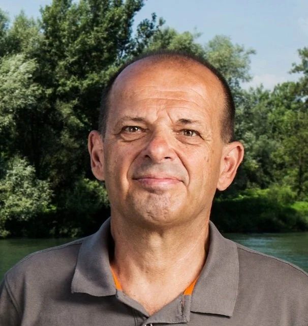 A middle-aged man with short brown hair and a slight smile, wearing a gray collared shirt, standing outdoors near a river with green trees in the background.