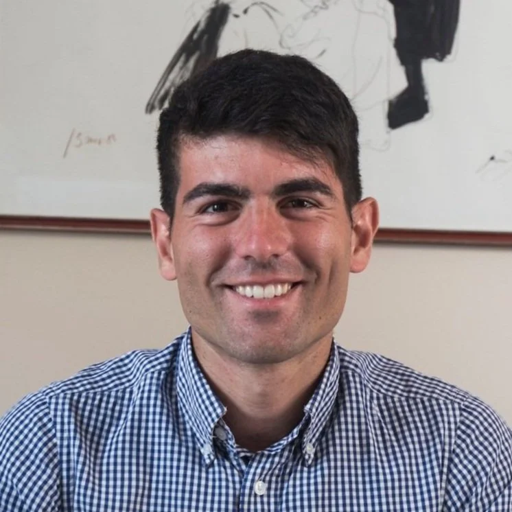 A man with short dark hair, smiling, wearing a blue and white checkered shirt, sitting in front of a whiteboard with abstract black markings.