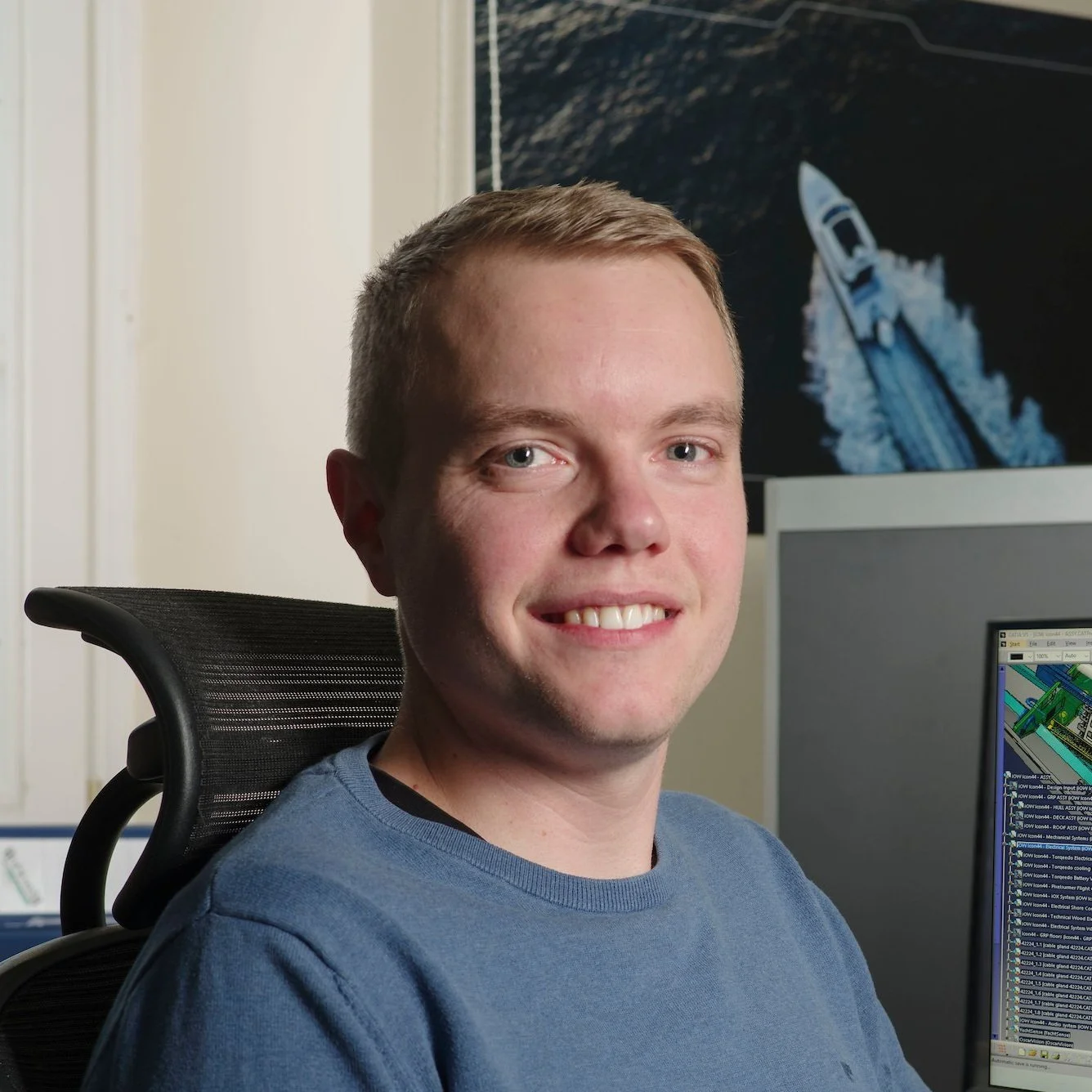 A young man with short blonde hair and blue eyes smiling at the camera, sitting in an office chair in front of a computer with a monitor displaying a colorful diagram or chart. In the background, there is a large framed photograph of a boat speeding through water.