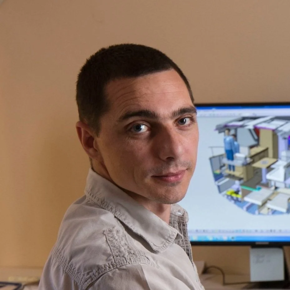 A man with short dark hair looking at the camera, in front of a computer monitor displaying a 3D design or architectural rendering.