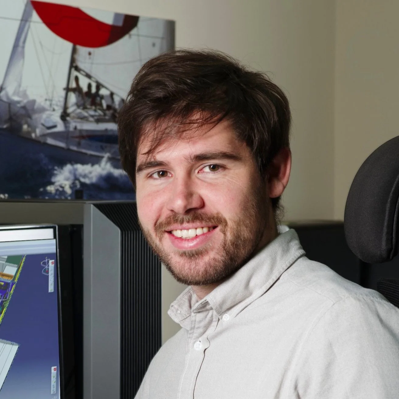 A man smiling sitting at a desk with computer monitors, with a large computer monitor and painting of a sailboat in the background.