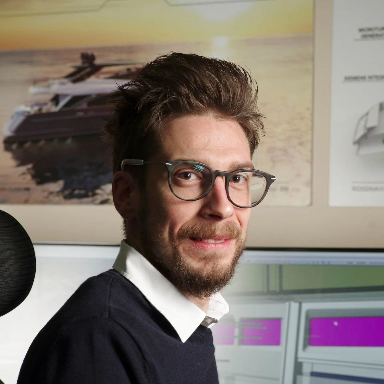 A man with glasses and a beard smiling at the camera in an office with posters and computer screens in the background.