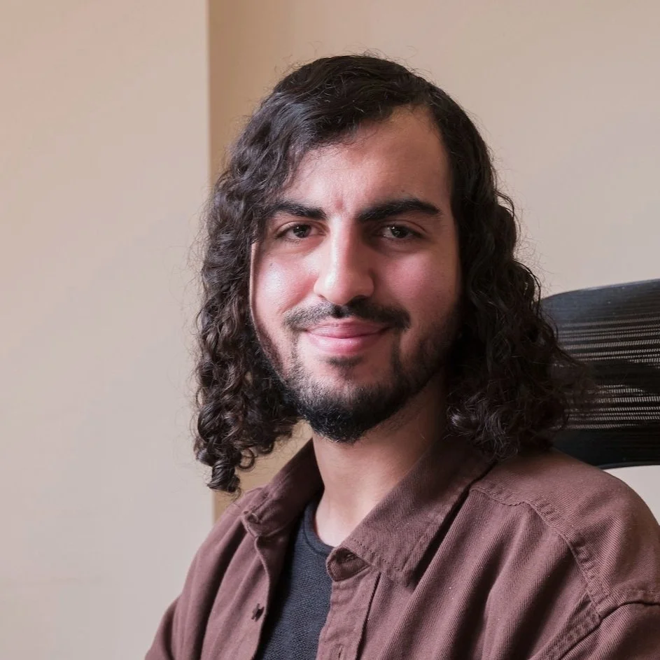 A man with long, curly dark hair and a beard sitting in a black office chair, smiling at the camera, wearing a brown shirt over a black t-shirt, against a neutral-colored wall background.