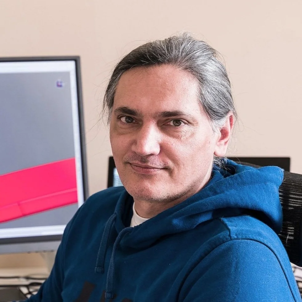 A man with shoulder-length gray hair sitting at a desk in front of a computer, wearing a blue hoodie, in an office setting.