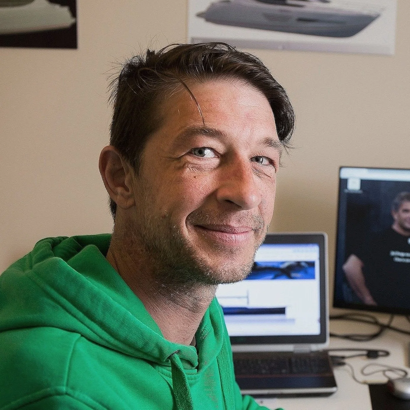 A man with short dark hair and a slight smile, wearing a green hoodie, sitting in front of a desk with two computer monitors, one displaying a woman.