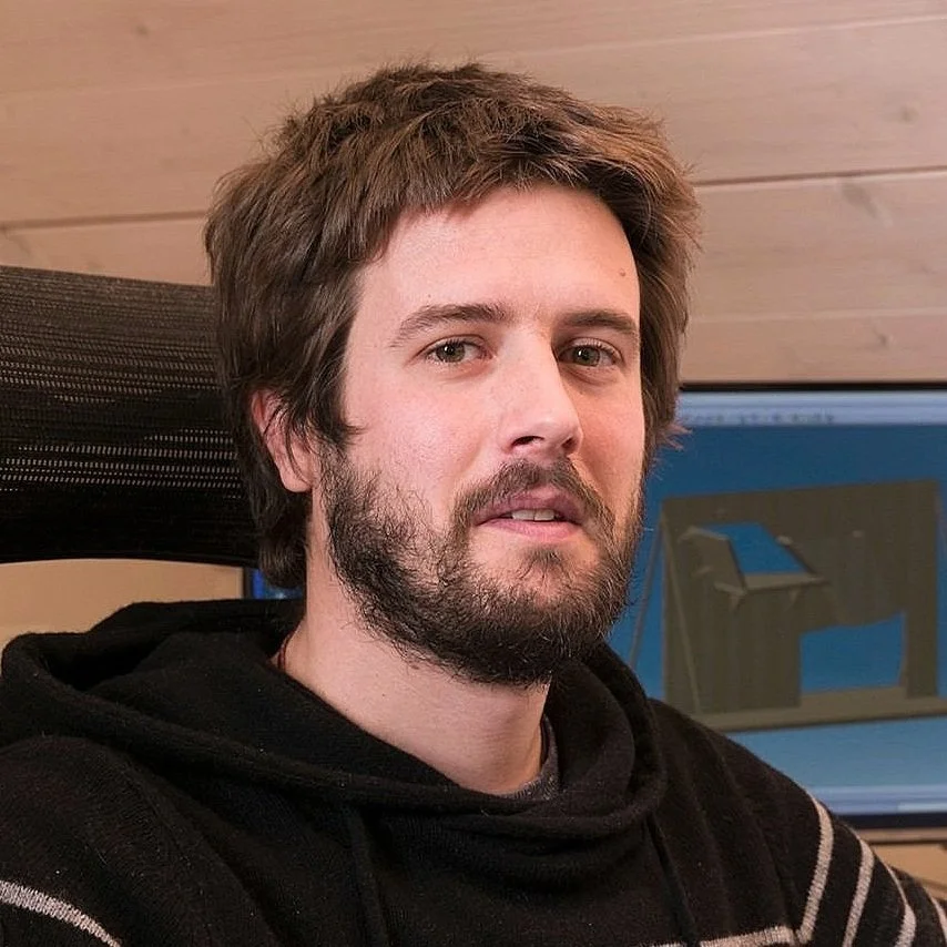 A young man with brown hair and a beard sitting in front of a computer monitor, wearing a black hoodie with white stripes, in a room with wood paneling on the wall.