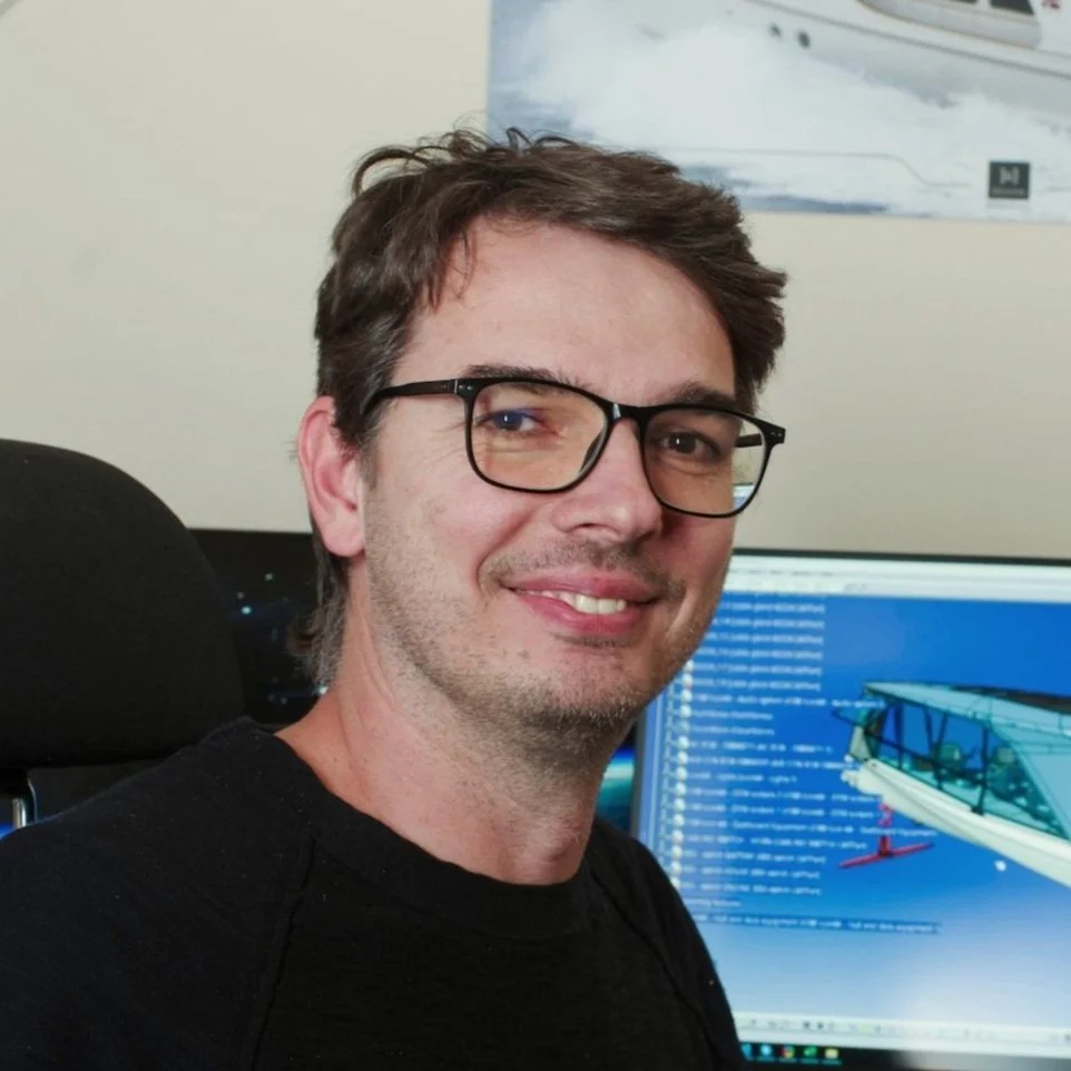 A man with glasses smiling in an office, with a computer monitor displaying a 3D model of a boat in the background.