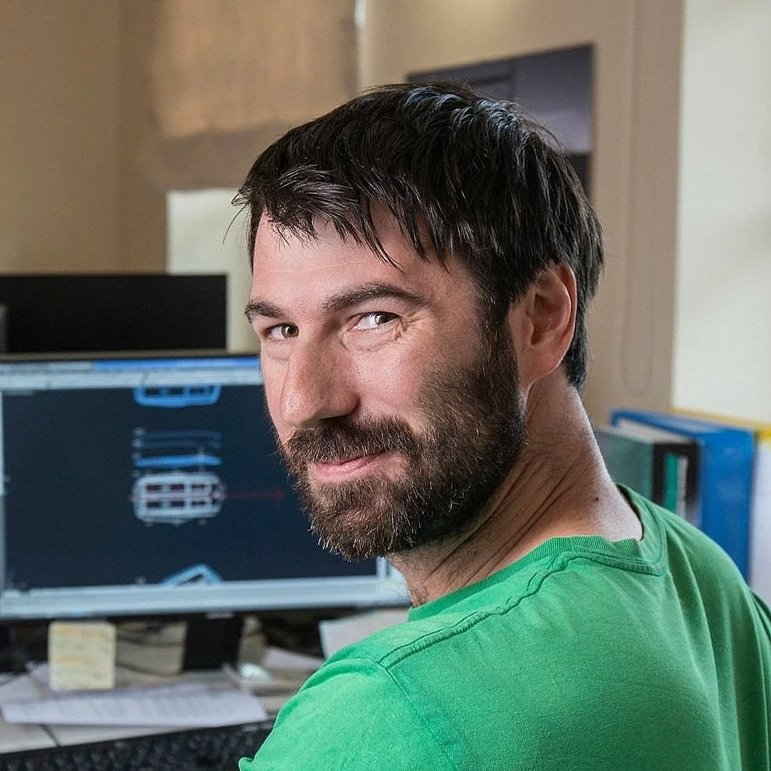 A man with dark hair and a beard, wearing a green shirt, sitting at a desk in front of monitors with technical drawings displayed on the screens.