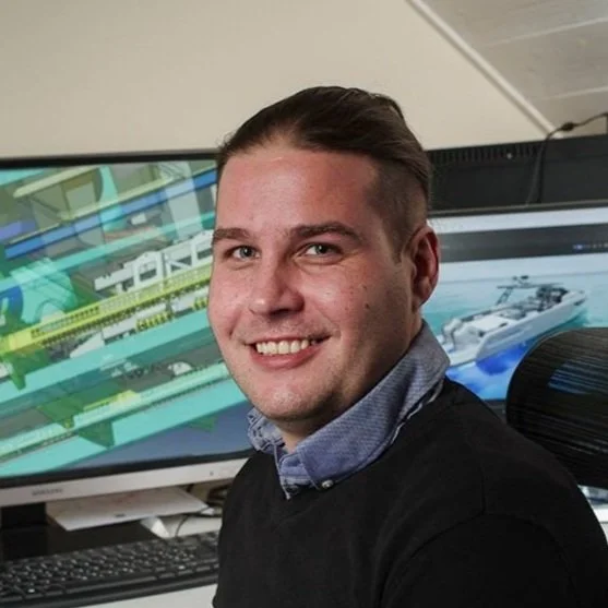 A young man with short dark hair smiling at the camera in front of a computer monitor displaying a technical or engineering design.