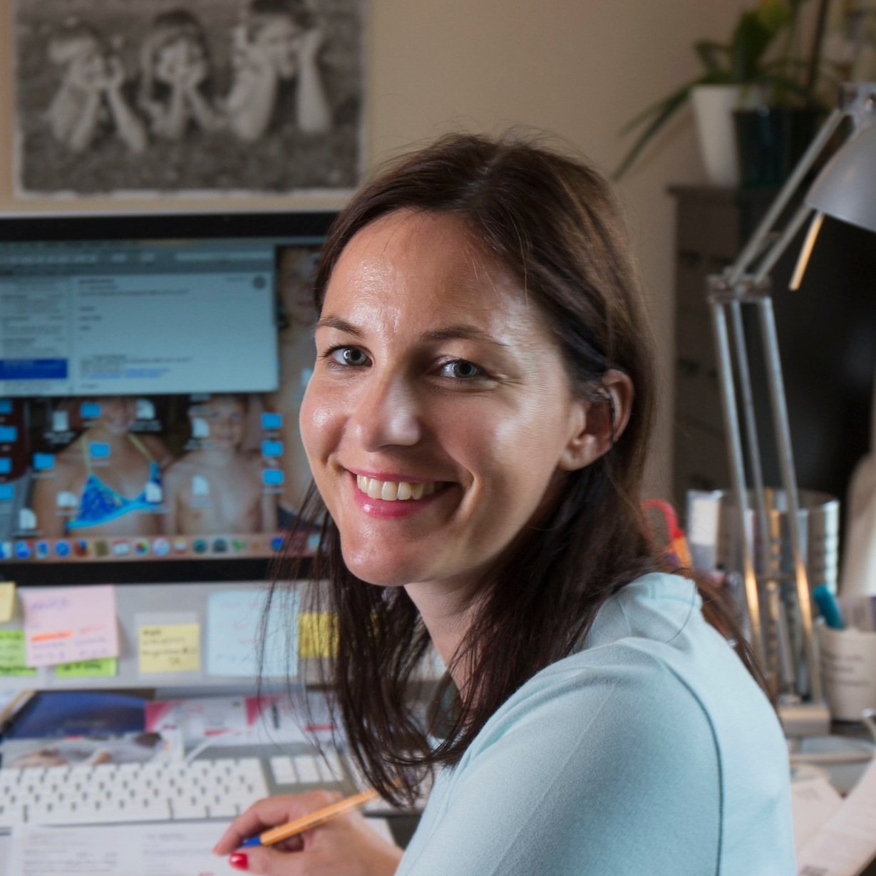 A woman with dark hair smiling at the camera in an office setting. There is a computer monitor and various office supplies in the background.