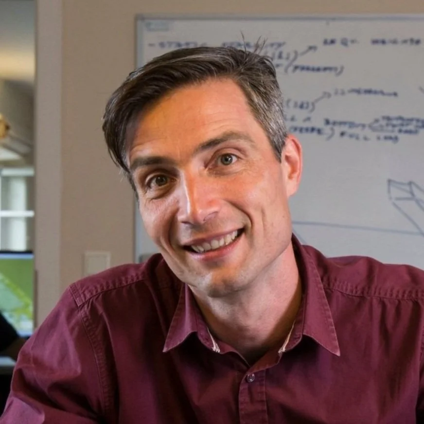 A man with brown hair and light skin smiling at the camera, wearing a maroon shirt, with a whiteboard with notes and diagrams in the background.