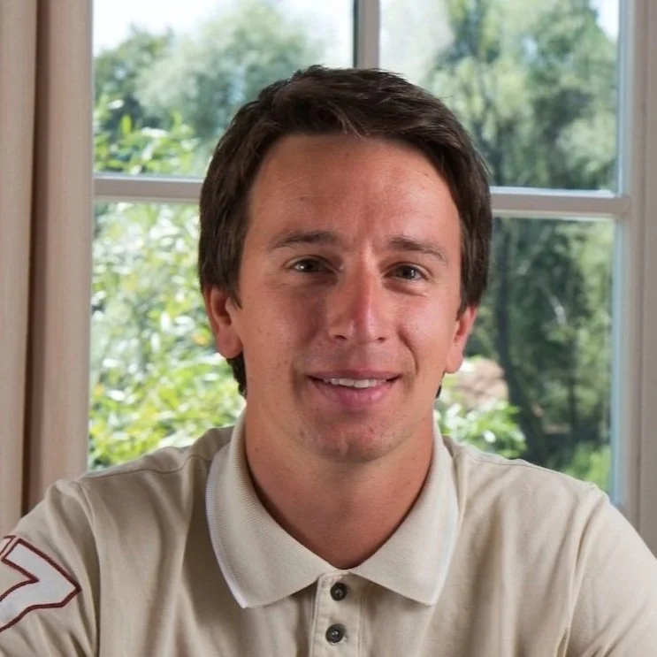 A young man with dark hair sitting indoors, smiling at the camera, in front of a window with greenery outside.
