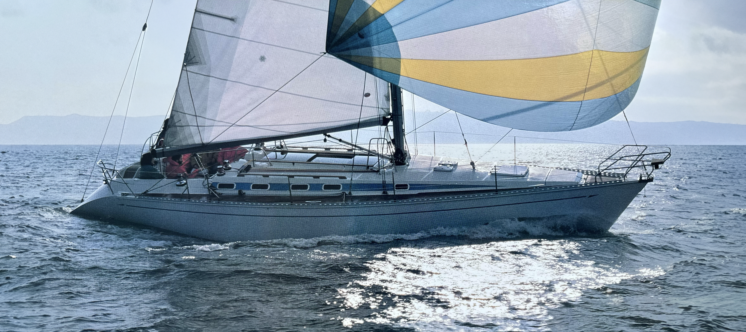 Sailboat with colorful spinnaker sailing on the ocean, distant land in the background, bright sunlight reflecting on water.