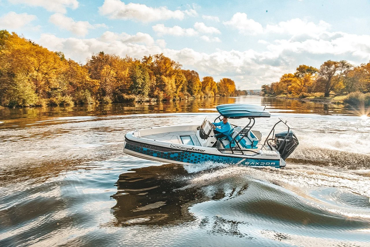 A person in a blue jacket driving a blue and white boat on a river with trees showing fall foliage on either side and a cloudy sky.