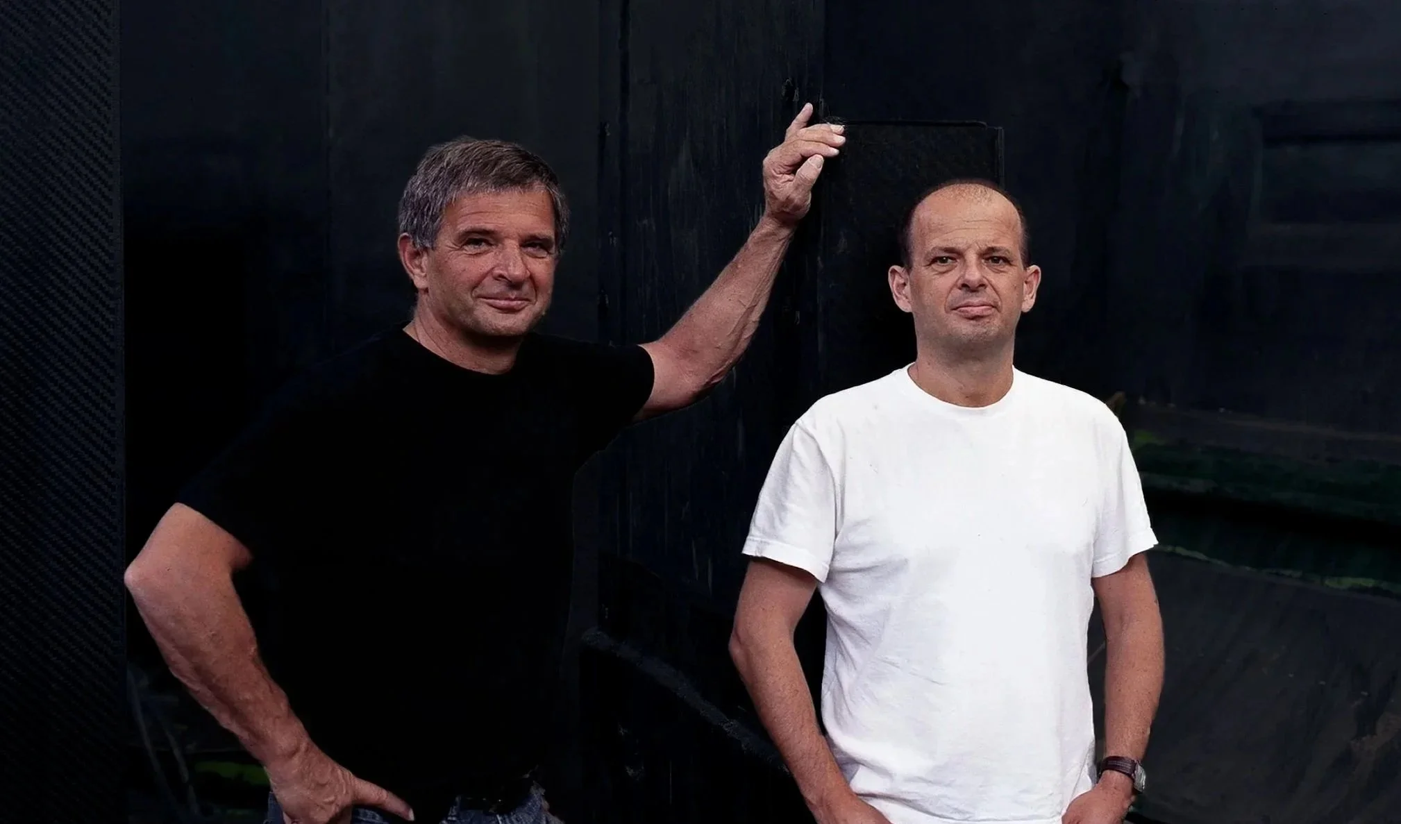 Two middle-aged men stand side by side in front of a dark background, one wearing a black shirt and the other wearing a white shirt. The man in black is touching a black surface with his arm extended.