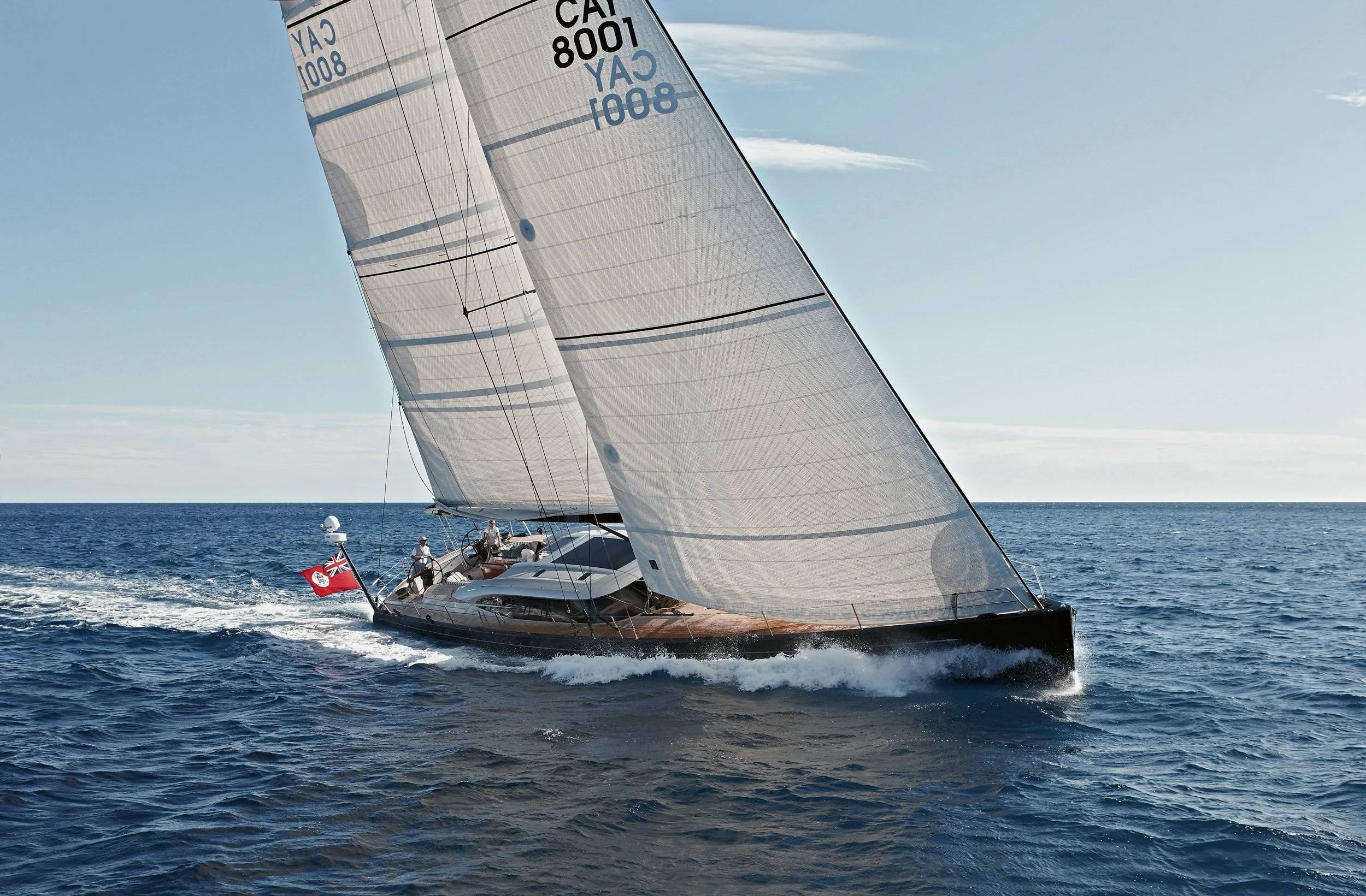 A luxury sailboat with large white sails on the open ocean, with a red flag at the stern and a clear blue sky.