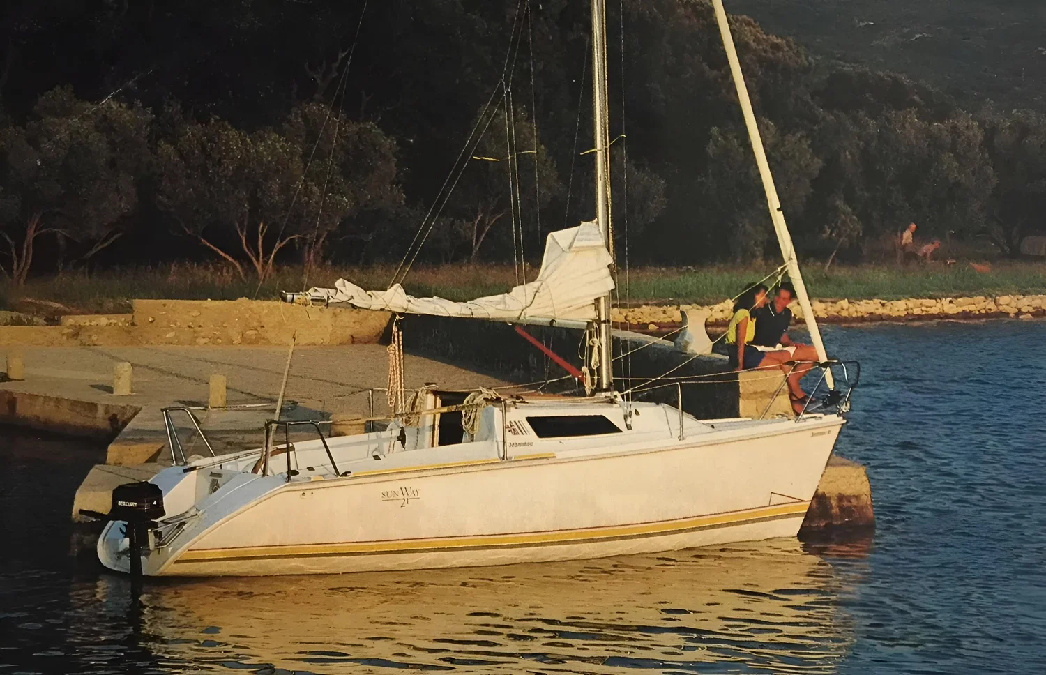 A sailboat docked at a harbor with two people sitting on the bow, during sunset with trees in the background.