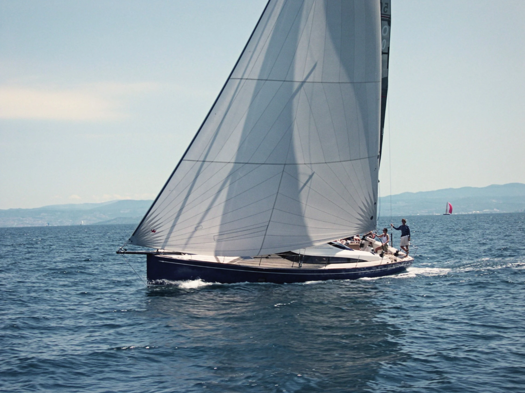A sailboat on the water with its sails hoisted, crew members on board, and distant land visible on the horizon.