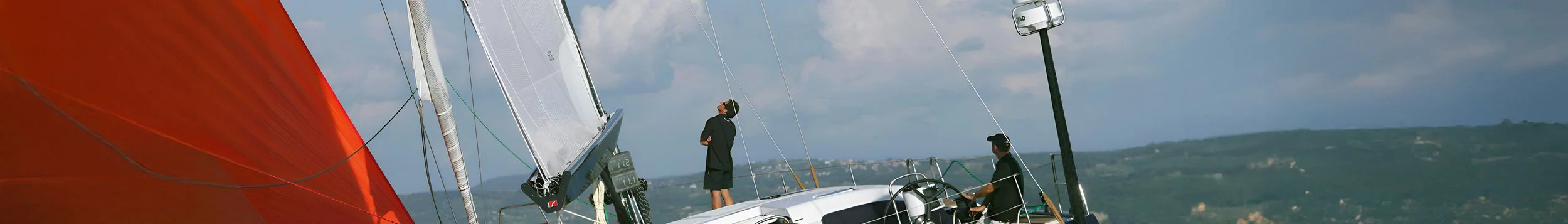 Two men on a sailboat with a red sail, navigating in open water with hills in the background, under cloudy skies.