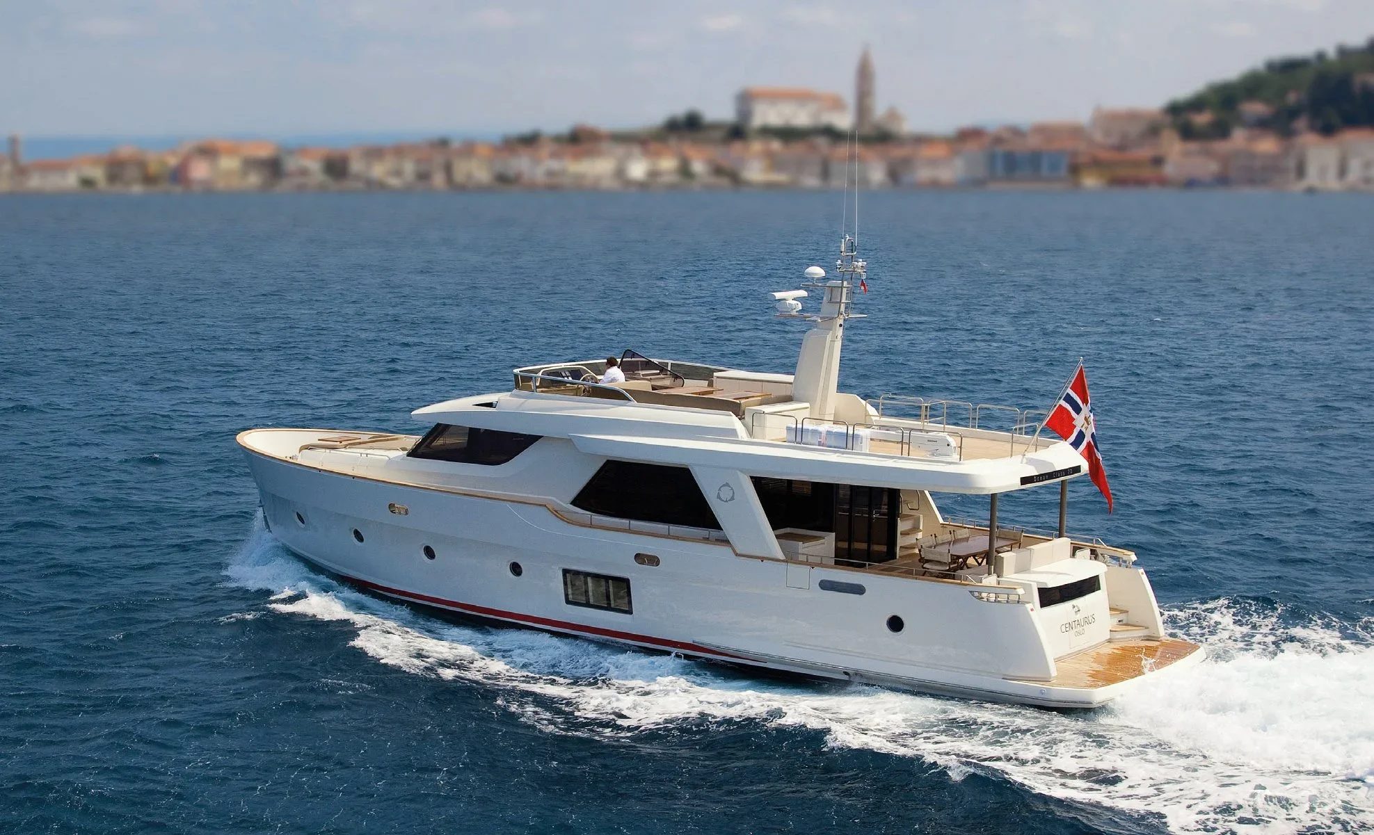 A white luxury yacht cruising on blue water near a coastal town with buildings and a church tower in the background.