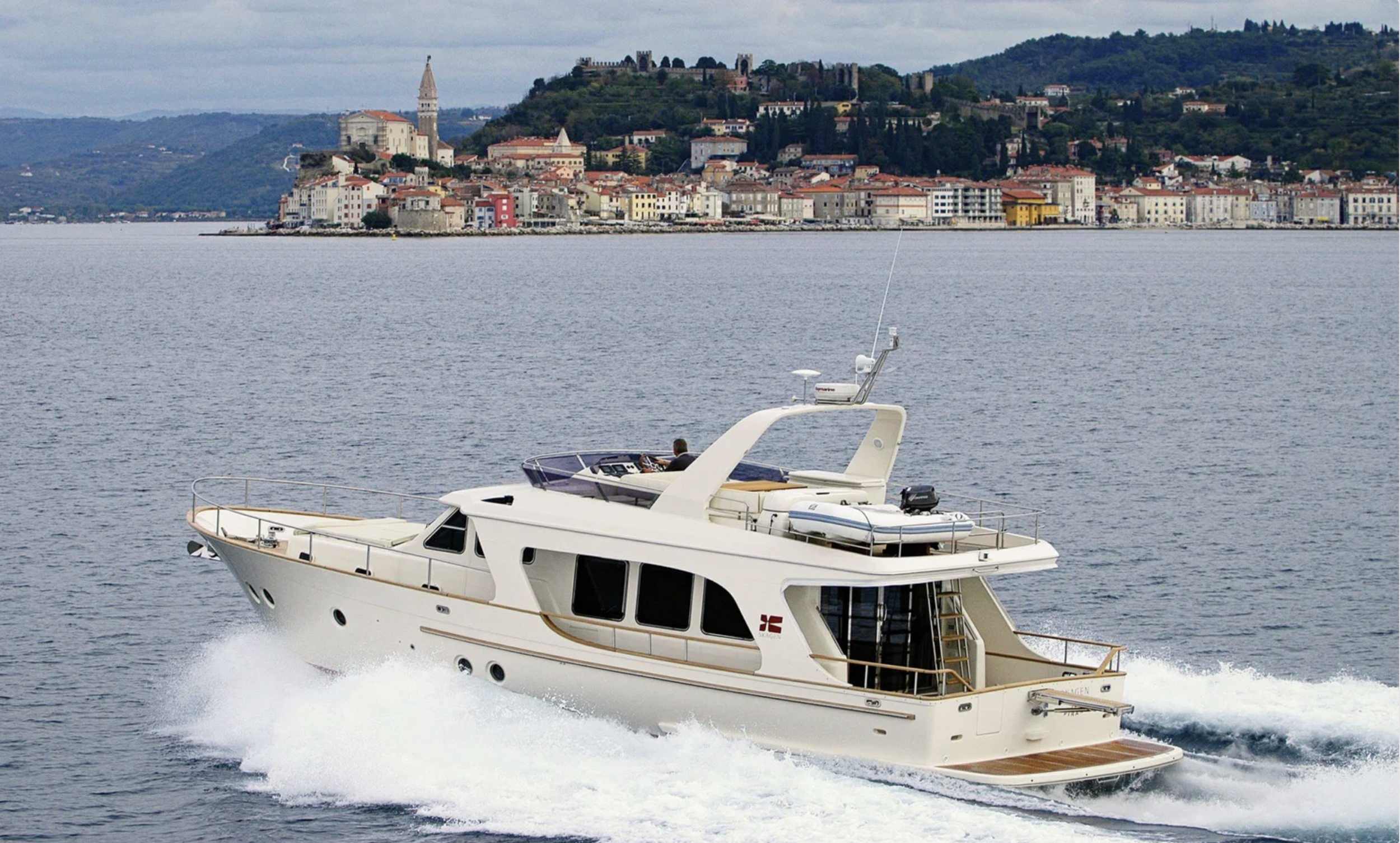 A white yacht sailing on a body of water with a scenic cityscape and hillside in the background.