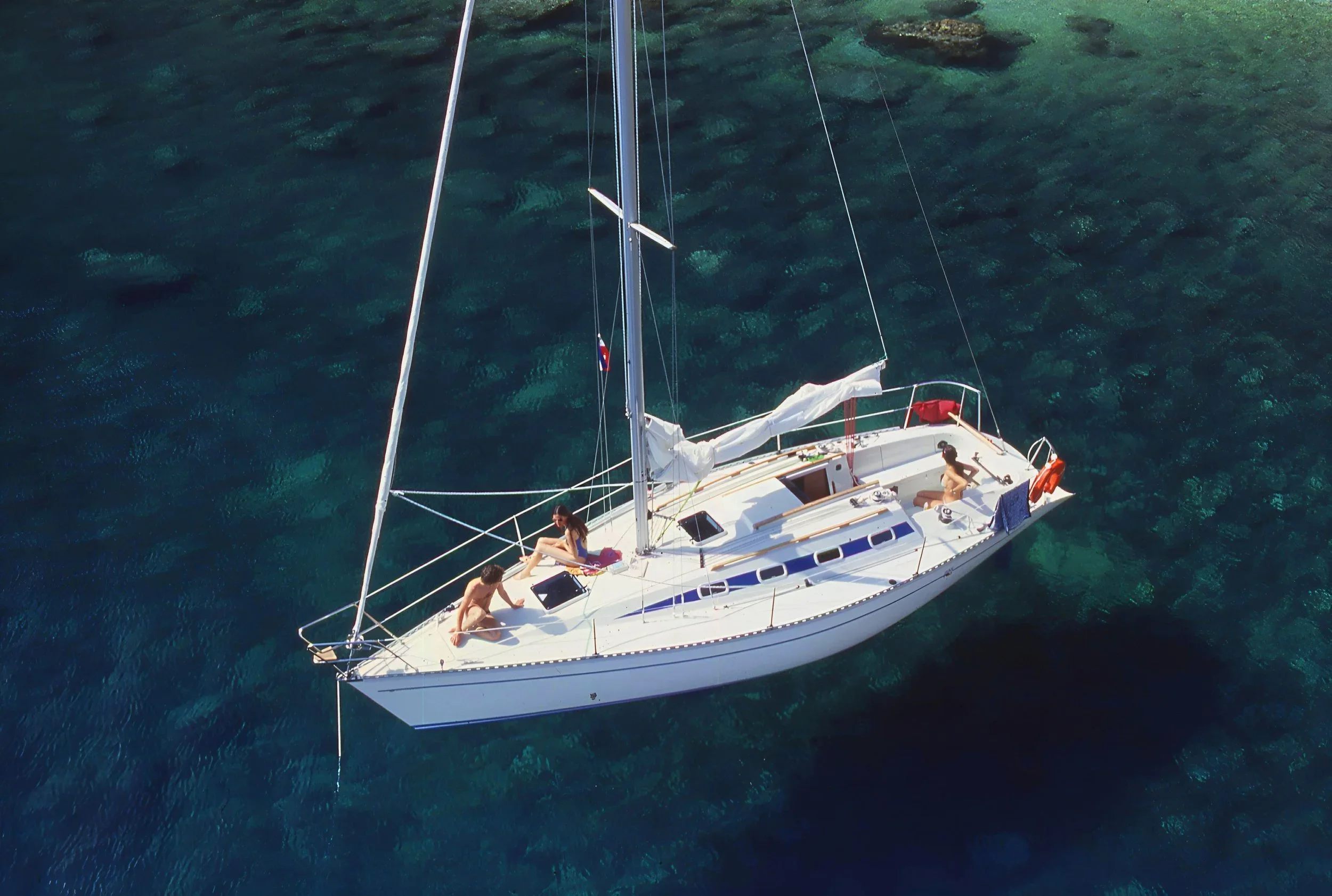 An aerial view of a white sailboat on clear blue water with three people on board, relaxing.