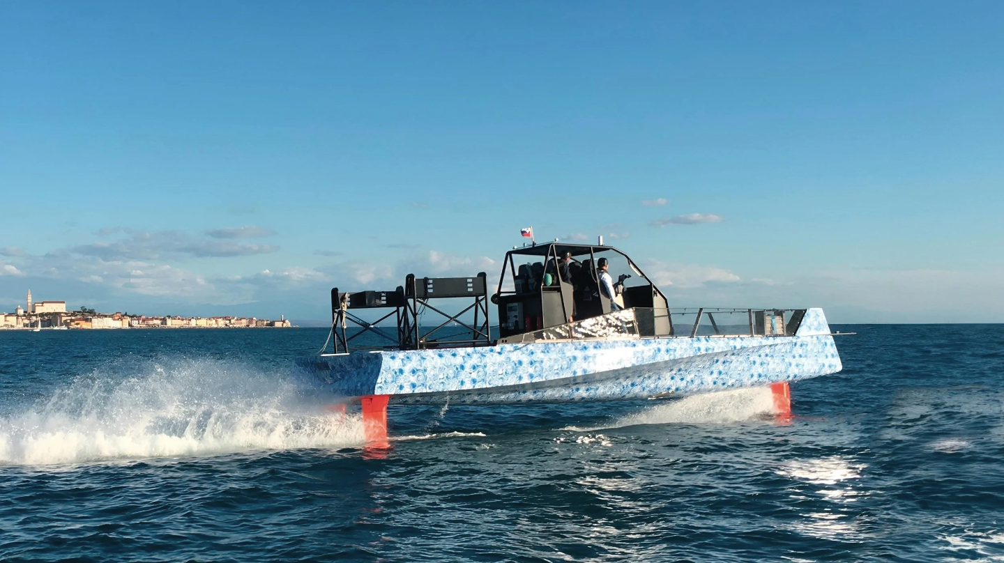 A futuristic hovercraft with a blue and white patterned exterior speeding across the water, with a city skyline in the background and a clear blue sky above.