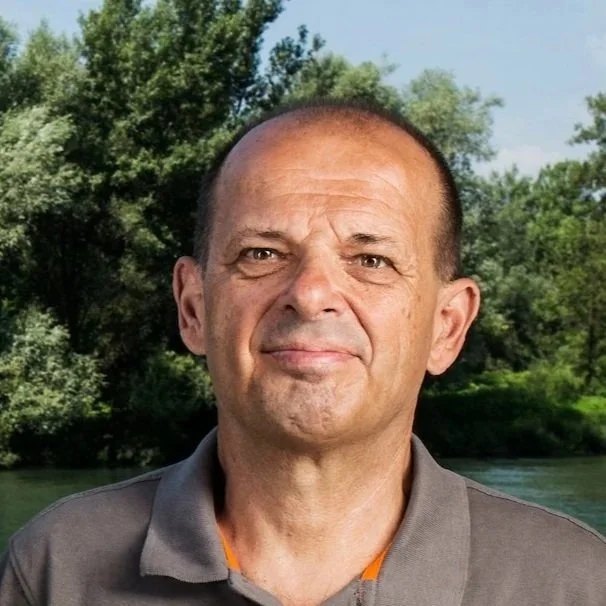 Close-up of a middle-aged man with a bald head and a slight smile, outdoors near a river and surrounded by green trees.