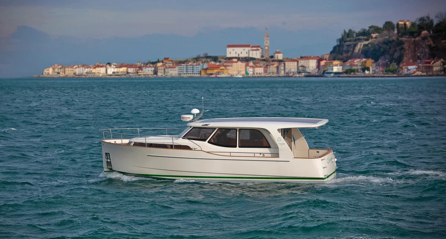 A white boat sailing on a blue body of water with a coastal town and buildings in the background.