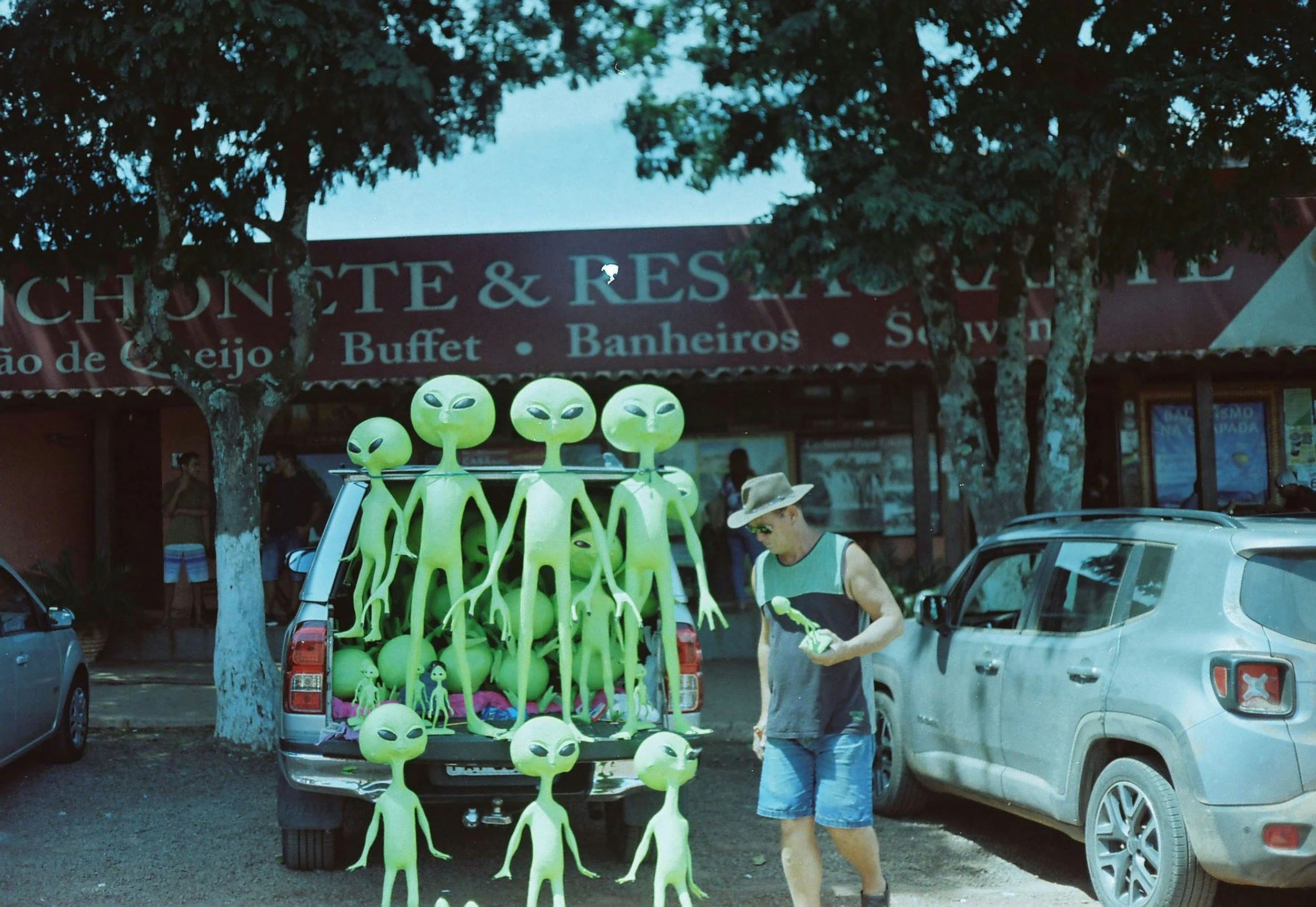 A man in a sleeveless shirt, shorts, sunglasses, and a wide-brimmed hat is standing next to a truck loaded with green alien plush toys, with some toys on the ground nearby, parked in front of a building with trees and cars around.