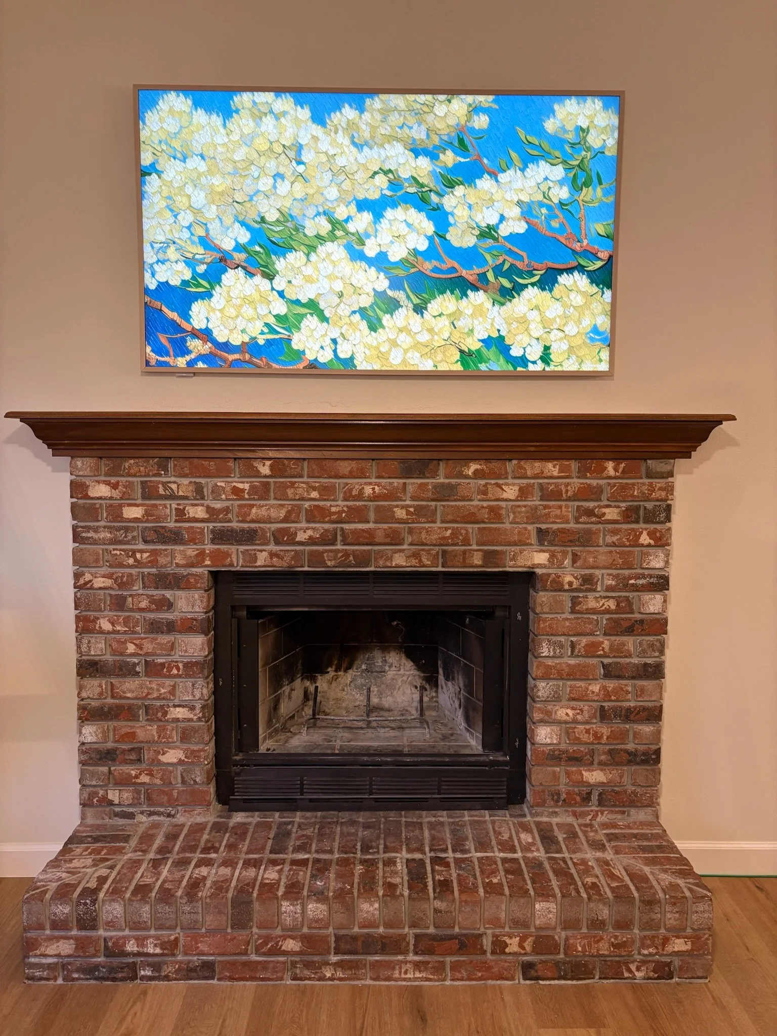 A brick fireplace with a wooden mantle and an empty metal firebox, topped with a large framed painting of white flowers on a blue background.