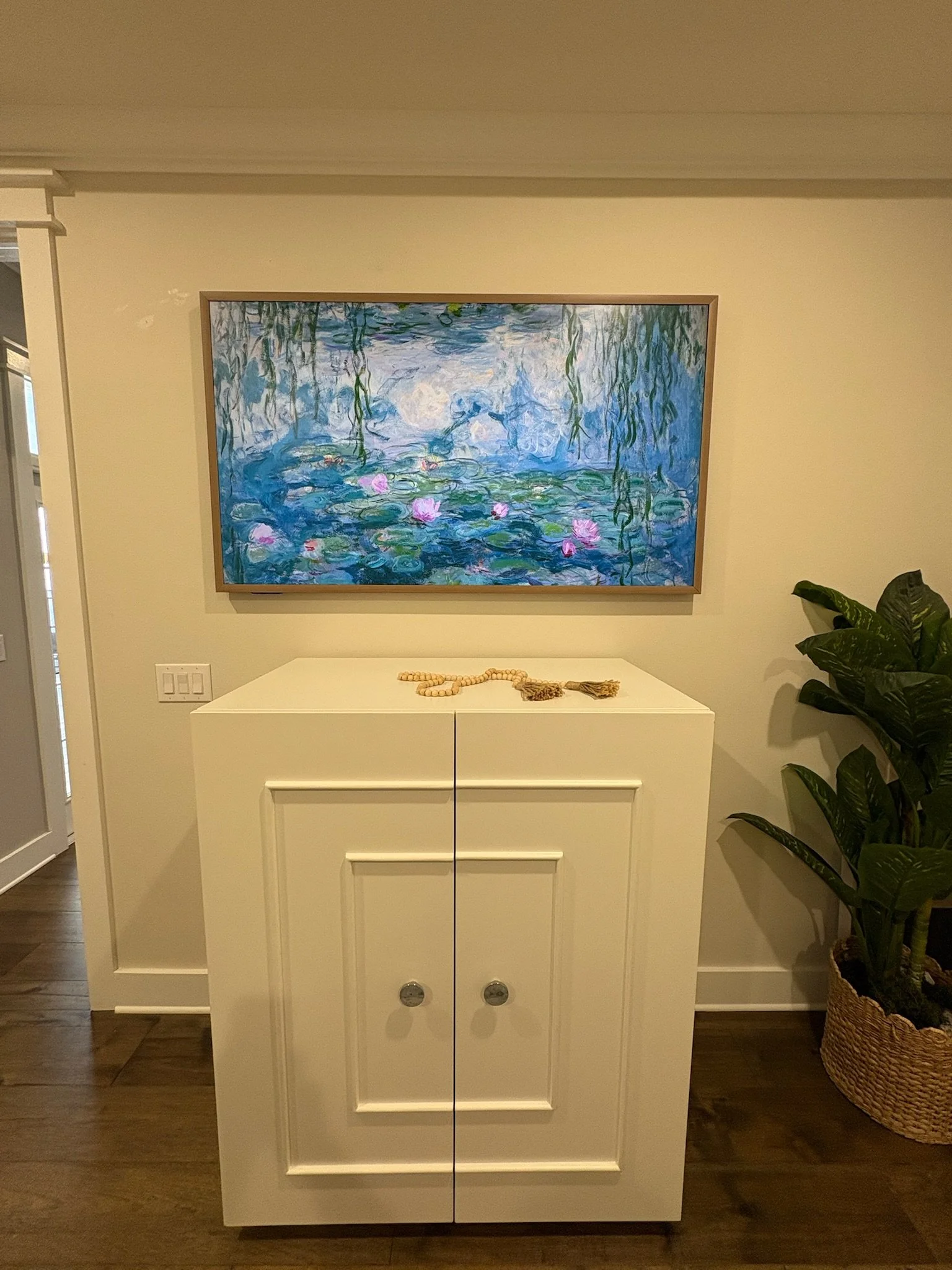 A white cabinet with two doors and silver knobs, placed against a beige wall. On top of the cabinet, there is a string of wooden beads with a tassel. To the right, a large leafy plant is in a basket. A framed impressionist style painting of a water l