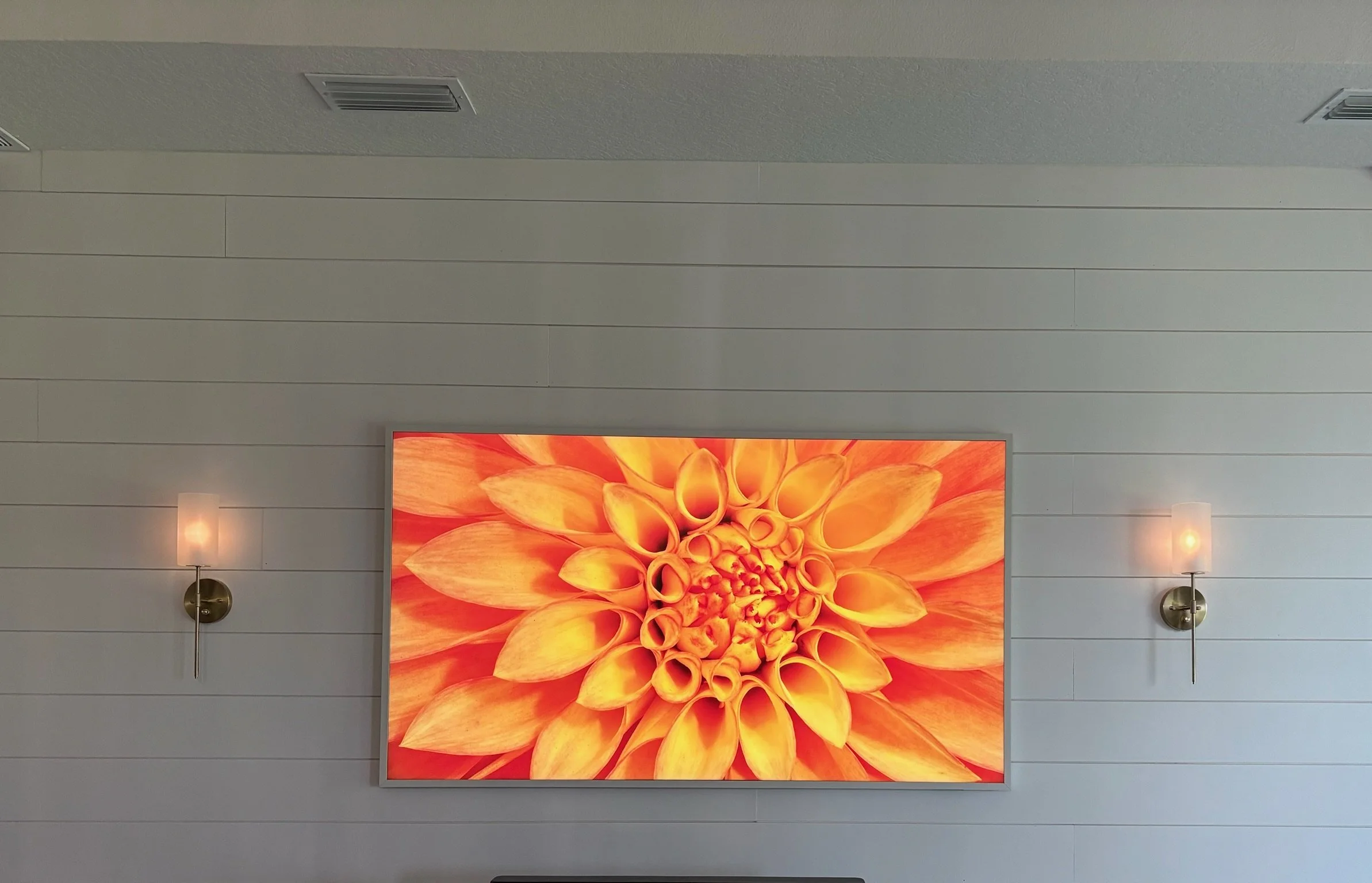 A flat-screen monitor displaying a close-up image of a vibrant orange flower with a detailed petal pattern, mounted on a light-colored wall with horizontal paneling and two wall-mounted lamps.