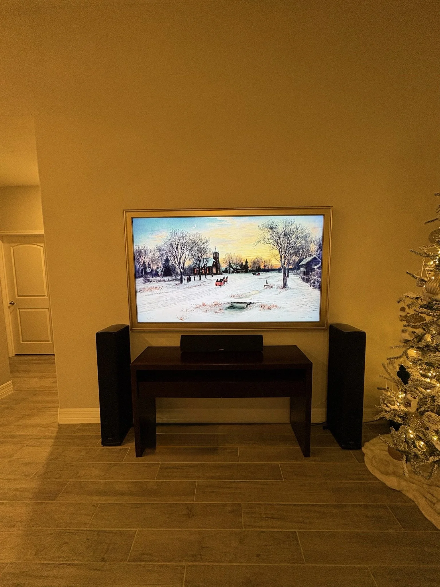 Living room with a TV displaying a winter scene with snow-covered trees and houses, a dark wooden console table, black speakers, a decorated Christmas tree on the right, and a closed white door with a curved panel on the left.