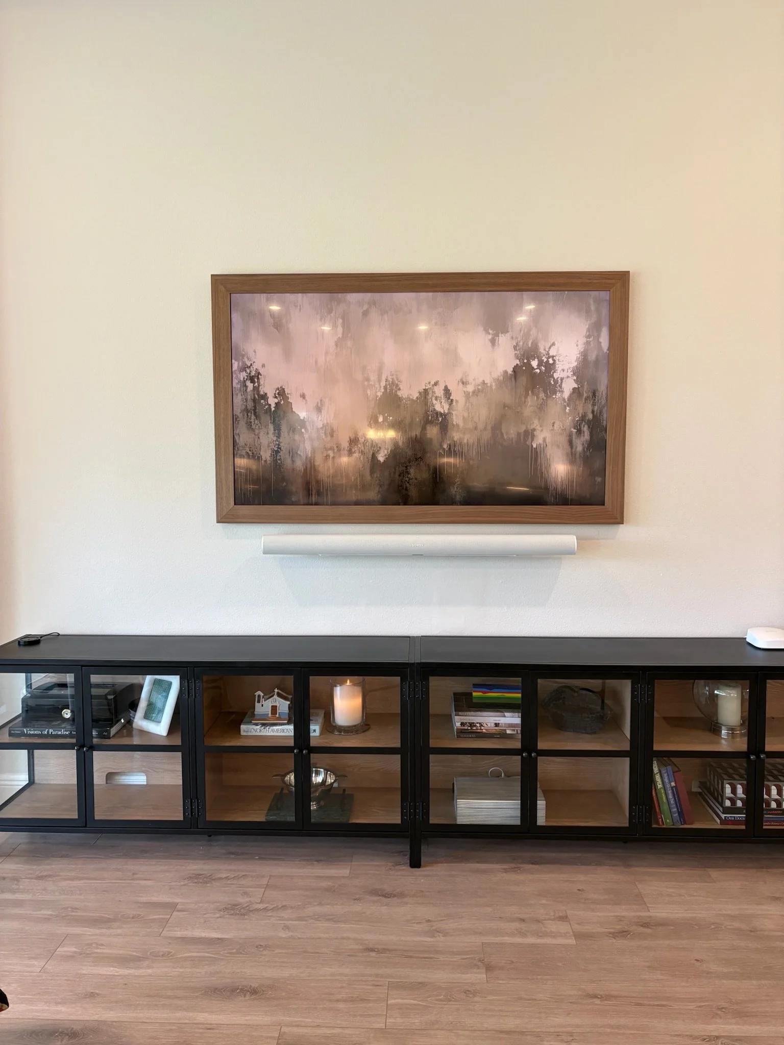 A black storage cabinet with glass doors, containing books, decorations, and candles, placed against a beige wall with a framed picture above it.