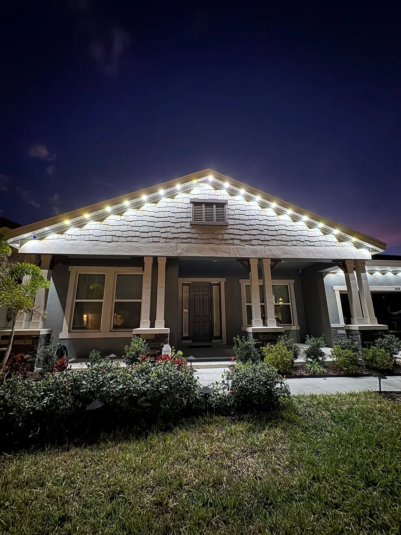 Night view of a house with a dark blue sky, illuminated by exterior lighting, featuring a front porch, landscaping, and a driveway.