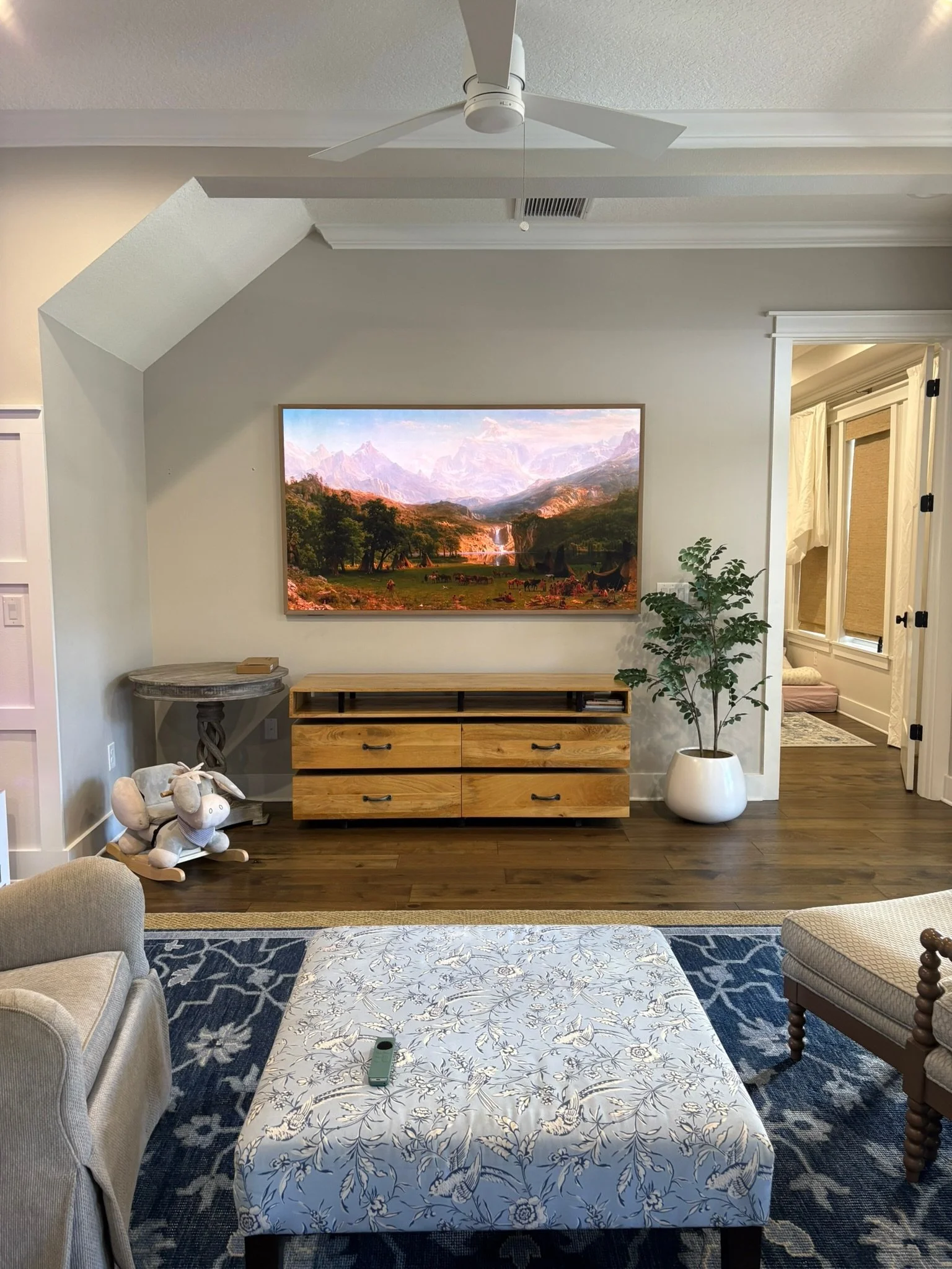 Living room with a large framed landscape painting, a wooden TV stand, a potted plant, a table with a blue and white patterned tablecloth, and an upholstered chair, with a doorway leading to a sunny corner with a window and curtains.
