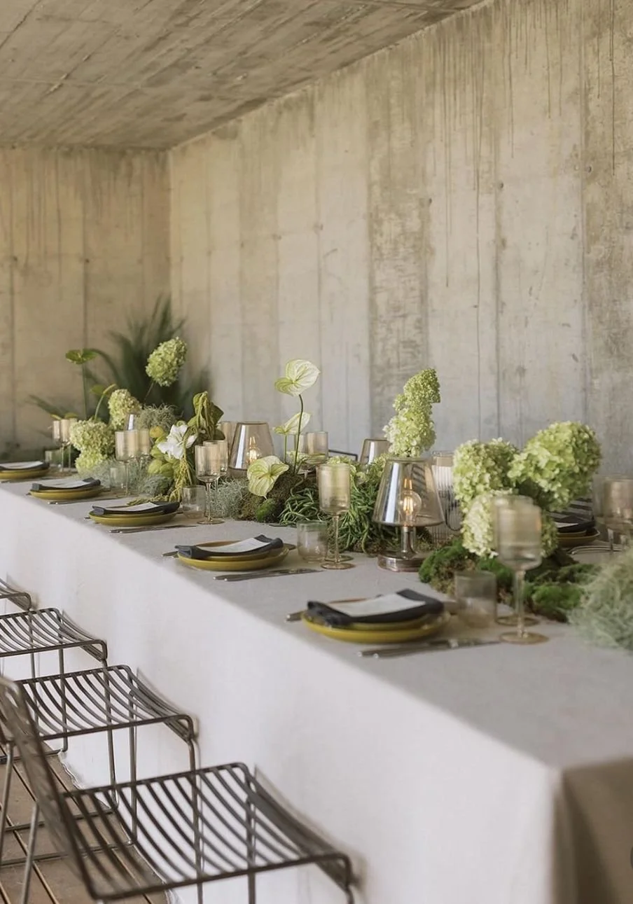 A long dining table decorated with green and white floral arrangements, candles, and place settings with plates, glassware, and napkins, set against a concrete wall.
