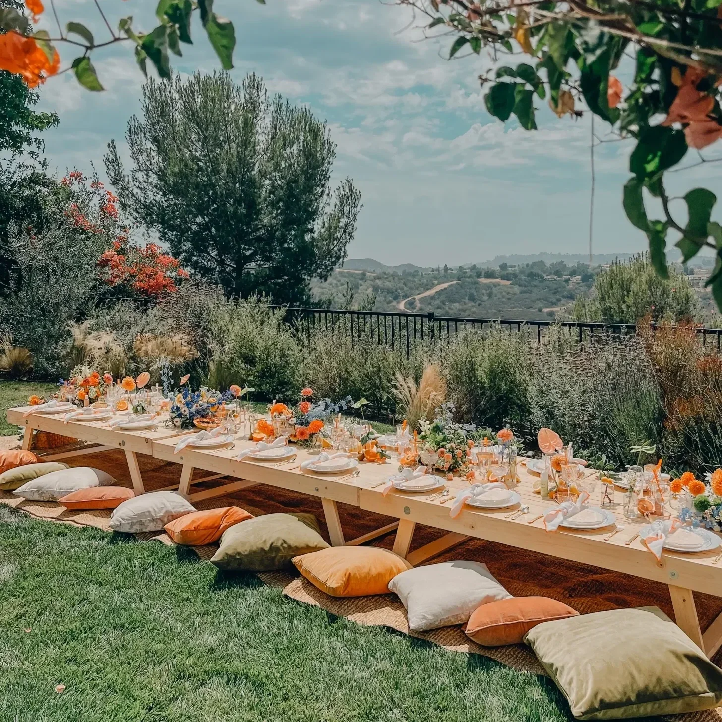 Outdoor long dining table decorated with orange and white flowers, set with white plates, napkins, and glassware, surrounded by colorful cushions on the grass, with a scenic view of hills and trees in the background.